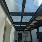 Pergola en métal avec toit en verre perforé, décorée de suspensions modernes et offrant un espace extérieur protégé et lumineux. Design contemporain pour une terrasse ou un jardin élégant.
