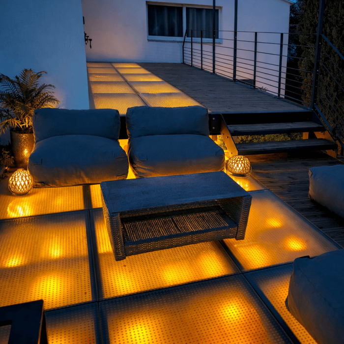 Terrasse moderne en verre éclairée de manière chaleureuse, avec des coussins confortables et une table basse, créant une ambiance accueillante pour un espace extérieur intime et lumineux.