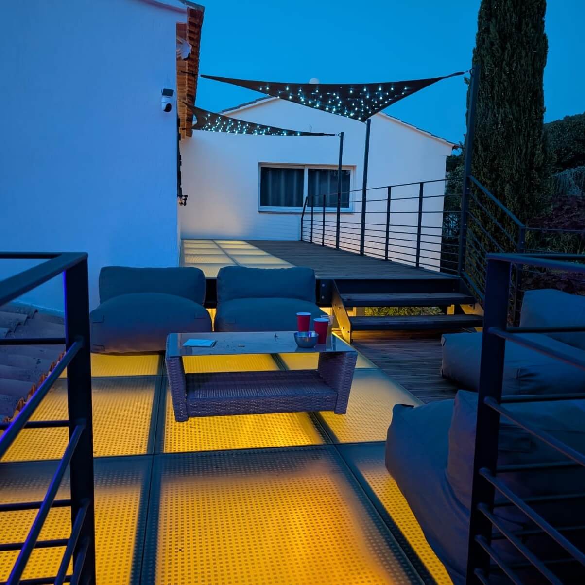 Pont en verre avec éclairage sous la surface, créant une ambiance moderne et élégante pour la terrasse, décoré de guirlandes lumineuses pour une atmosphère agréable en soirée.