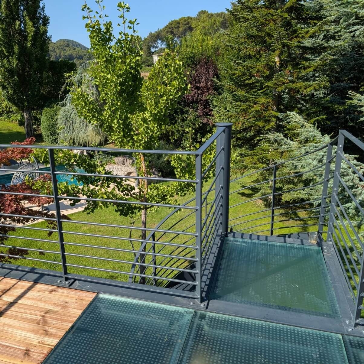 Pont en verre moderne avec garde-corps métallique, menant à une terrasse avec une vue dégagée sur un jardin luxuriant, entouré de verdure et d'une piscine.